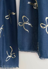 Sciarpa blu navy realizzata in tessuto strutturato con motivi di fiocchi a nastro dorati. I bordi sono sfrangiati per un dettaglio in più e un interesse visivo.