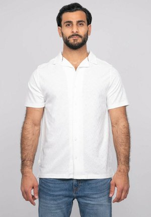 Homme avec une barbe portant une chemise blanche texturée à manches courtes boutonnée et un jean bleu, debout devant un fond clair uni.