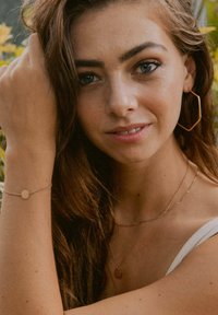 Schmuck aus Roségold mit einem kreisförmigen Armband und geschichteten Halsketten, dazu ein sechseckiger Ohrring. Das Modell hat lange, wellige braune Haare.