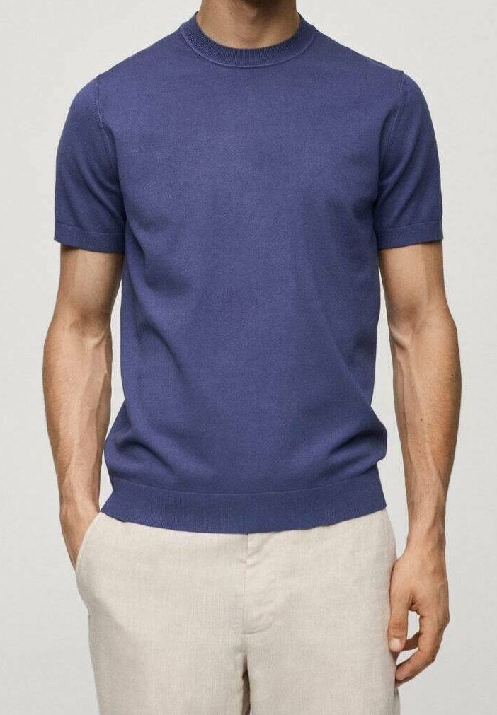 Homme portant un pull bleu à manches courtes et col rond, associé à un pantalon beige clair, debout avec une main dans la poche.
