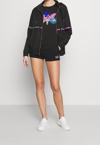 Person med svart zip-up jacka, rosa och blå logot-shirt, svarta shorts med vit logotyp och vita sneakers, står mot en enfärgad vägg.