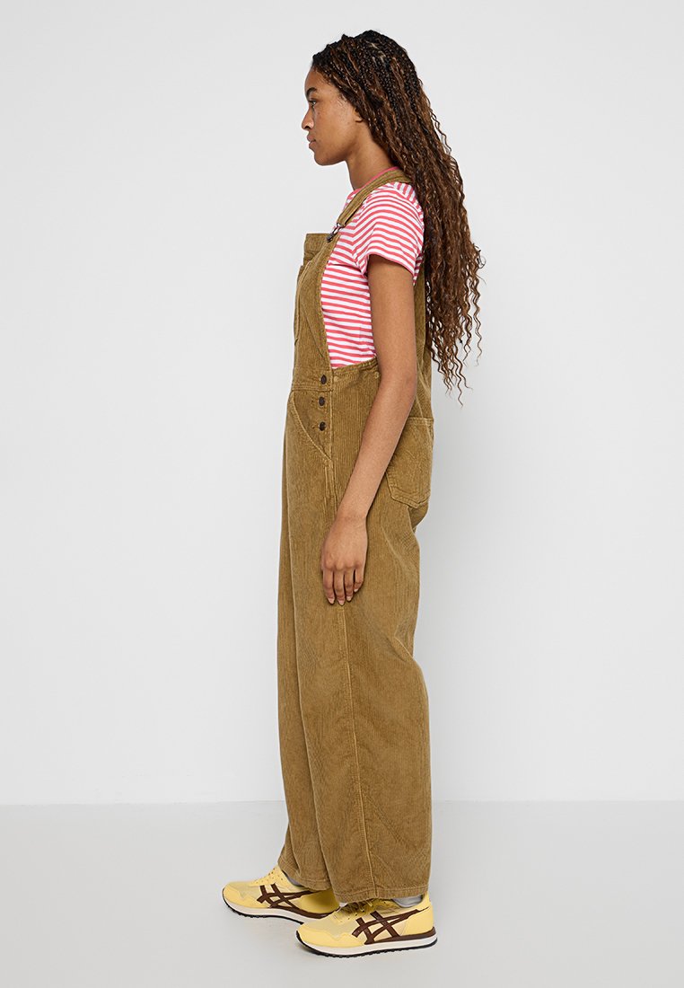 Salopette en velours côtelé marron avec une coupe ample, des poches latérales et des détails de boutons, associée à une chemise rayée rouge et blanche et des baskets jaunes.
