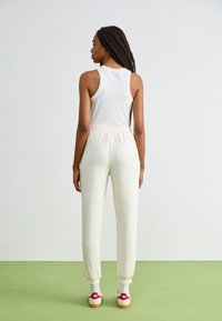 Witte geribbelde tanktop en lichtbeige joggers met een elastische tailleband, gecombineerd met sneakers met rode accenten, staande op groene vloer.