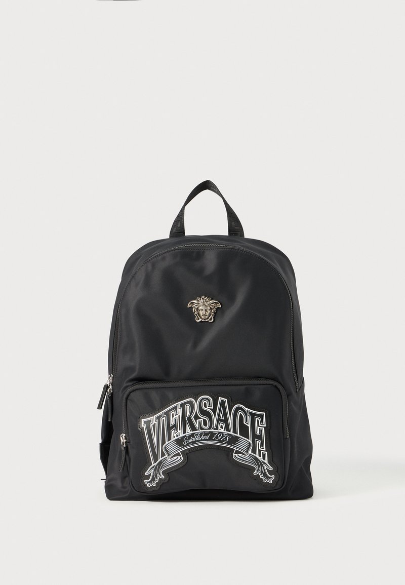 Rucsac negru din nylon, cu un logo aurit Medusa proeminent, buzunar frontal cu fermoar și detalii brodate "VERSACE" în alb. Textură netedă și formă rotunjită.