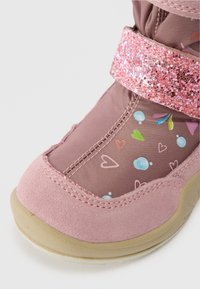 Közeli kép egy rózsaszín gyermekcipőről, csillogó pánttal, szív- és buborékmintákkal, valamint bézs talppal, fehér háttéren.