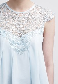 Vaaleansininen hihaton paita, jossa on yksityiskohtainen pitsipäällyste kaula-aukossa. Paita on valmistettu virtaavasta, läpikuultavasta kankaasta ja se istuu löysästi.