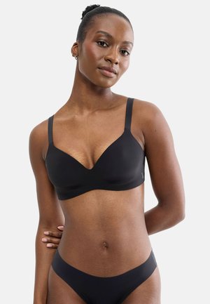 Woman with dark skin and hair in a bun wearing a black bra and matching black underwear against a plain light background.