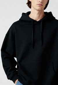 Sudadera negra de material de algodón, con bolsillo tipo canguro, capucha con cordones y puños acanalados. Diseño minimalista con un ajuste relajado.