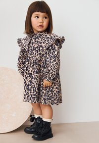 Leopardenmuster-Kleid mit Rüschkragen, langen Ärmeln und tailliertem Schnitt. Schwarze Schnürstiefel mit gerippten Socken. Der Hintergrund zeigt eine runde Form in heller Farbe.