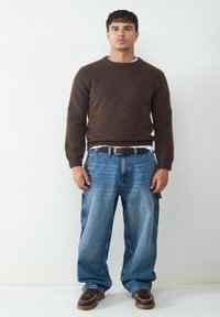Brauner Pullover mit gerippten Bündchen, lockere blaue Denim-Cargohosen und braune Lederschuhe mit Ziernähten, stehend vor einem hellen Hintergrund.