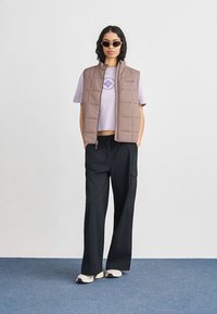 Γυναίκα που φοράει γυαλιά ηλίου, καφέ μπουφάν με επένδυση πάνω από λιλά μπλούζα, μαύρο παντελόνι με φαρδιά πόδια και λευκά αθλητικά παπούτσια, στέκεται σε μπλε χαλί.