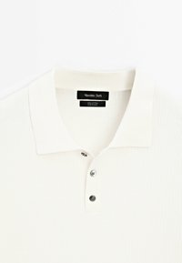 Polo en coton blanc côtelé avec trois boutons et un col pointu, étiqueté « Massimo Dutti » et fabriqué en Chine.