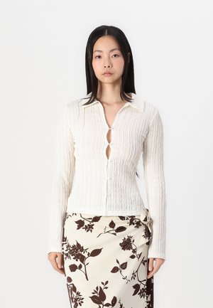 Long-sleeve white blouse with a textured pattern, featuring a button-down front and collar. Worn with a patterned brown and white skirt.