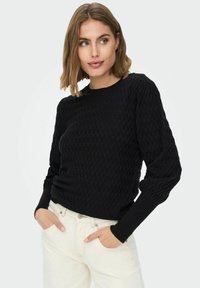 Svart teksturert genser med rund hals og ballong-ermer, kombinert med hvite jeans. Stoffet har et hevet bikube-mønster.