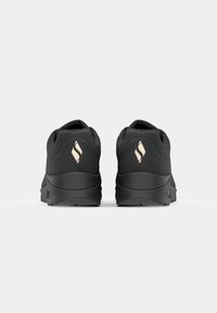 Paire de chaussures de sport noires vues de dos avec un logo blanc stylisé sur chaque talon.