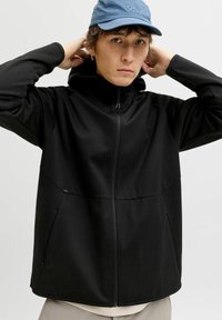 Veste noire zippée avec capuche, présentant une texture lisse et deux poches latérales. Le modèle porte une casquette bleu clair pour créer un contraste.