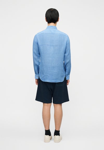 Ανοιχτό μπλε πουκάμισο με μακριά μανίκια από λινό ύφασμα, με ίσια γραμμή και μανσέτες με κουμπιά, συνδυασμένο με σκούρα σορτς και αθλητικά παπούτσια.
