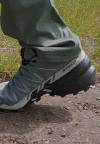 Chaussure de randonnée grise avec une surface texturée, semelle noire et accent blanc. La semelle présente des signes d'usure. Fond en herbe et terre.
