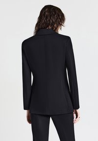 Blazer nero aderente con un design strutturato, maniche lunghe e colletto a pistagna. Tessuto liscio con silhouette sagomata sul retro.