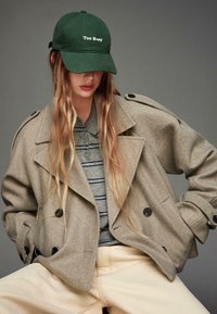 Căciulă de culoare verde măsliniu cu textul "Prea ocupat", tricou polo cu dungi gri, palton oversized din lână bej cu nasturi negri și pantaloni de culoare deschisă.