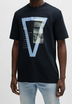 Mężczyzna noszący czarny t-shirt z krótkim rękawem z dużym niebieskim napisem „V7” oraz czarno-białymi zdjęciami gór i tekstem z przodu, w połączeniu z jasnoniebieskimi dżinsami.