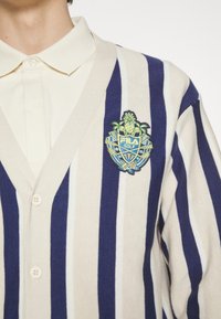 Gestreifter Strickcardigan in Marineblau und Creme mit vertikalen Streifen. Verfügt über einen V-Ausschnitt, einen Knopfverschluss und ein gesticktes FILA GOLF-Logo mit Ananas-Detail.