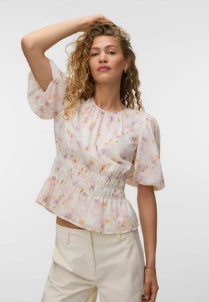Femme aux cheveux bouclés portant un chemisier léger à fleurs avec des manches bouffantes et un pantalon crème taille haute, posant avec une main sur la tête.
