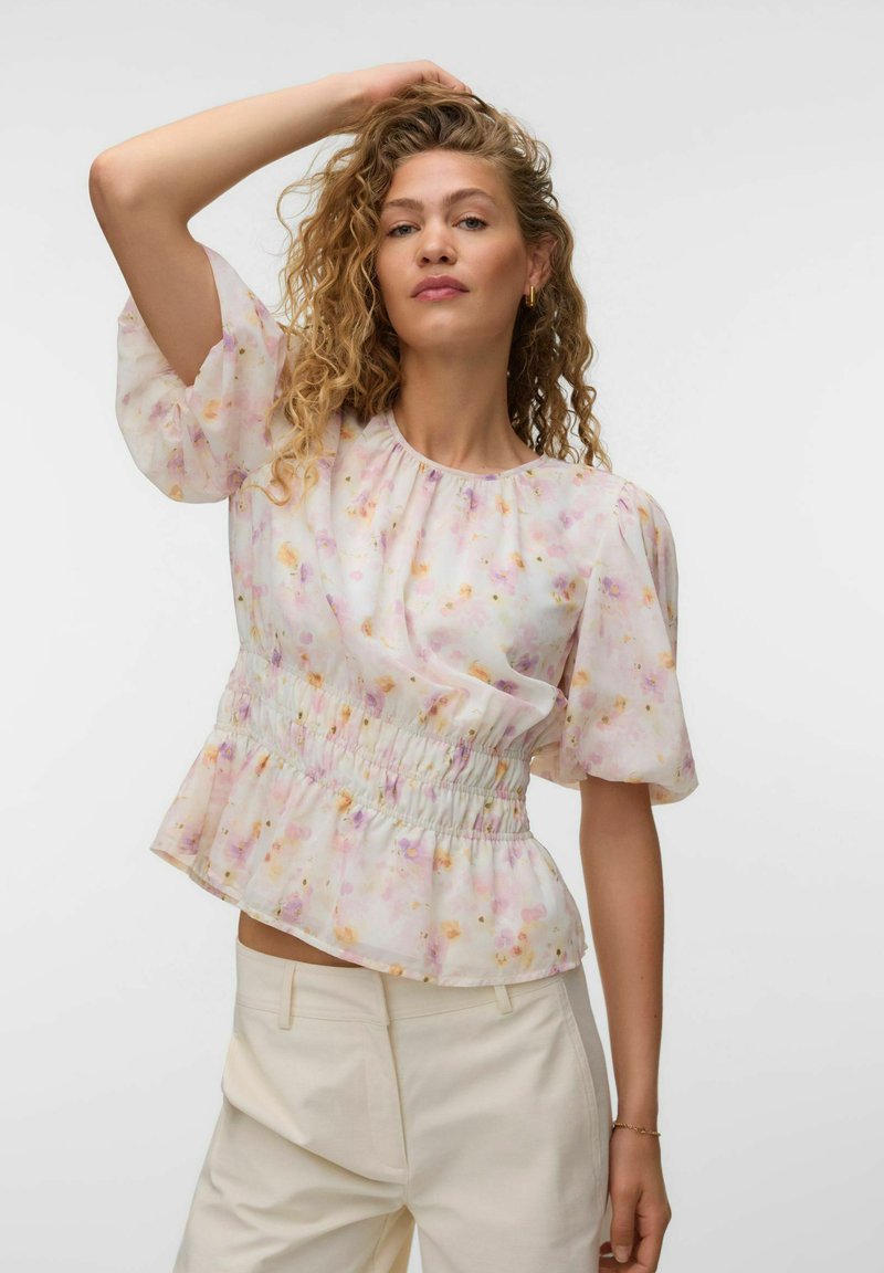 Femme aux cheveux bouclés portant un chemisier léger à fleurs avec des manches bouffantes et un pantalon crème taille haute, posant avec une main sur la tête.