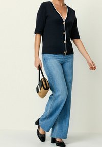 Navy cardigan with white trim and gold buttons, paired with light blue denim flared jeans. Black and straw crossbody bag and black heeled shoes.