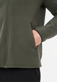 Olivgrüne Fleecejacke mit einem Reißverschluss vorne und zwei Seitentaschen, aus strukturiertem Stoff mit marineblauen Kordelzügen.