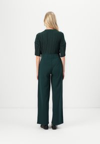 Zelena plisirana bluzica s kratkim rukavima u kombinaciji s hlačama širokih nogavica visokog struka, oboje od glatkog materijala, viđeno s leđa.