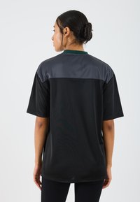 Svart sportsskjorte med grå paneler på skuldrene, korte ermer og rund hals. Stoffet ser glatt og lett ut.