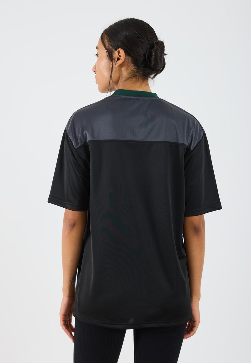 Camisa deportiva negra con un panel gris en los hombros, mangas cortas y un escote redondeado. La tela parece suave y ligera.