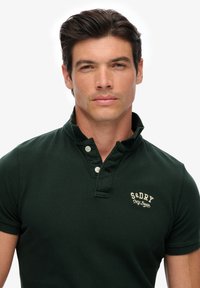 Donkergroen poloshirt van gestructureerde stof met een kraag, voorzien van drie witte knopen en een lichtbruin geborduurd logo op de borst.