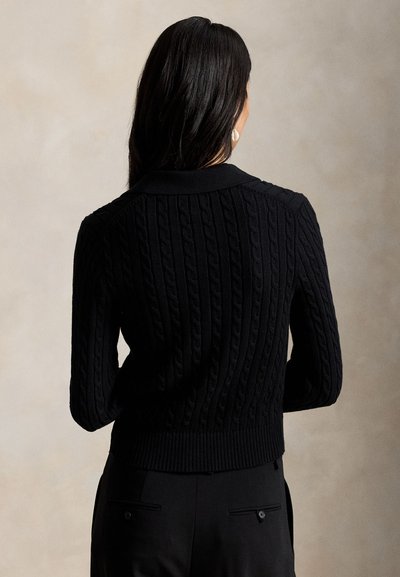 Kvinde med langt mørkt hår vendt væk, iført en sort kabelstrikket sweater og sorte bukser mod en ensfarvet beige baggrund.
