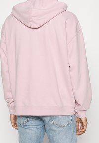Sudadera rosa claro de gran tamaño con capucha ajustable, puños y dobladillo acanalados. Combinada con jeans azul claro, que muestran una suave textura de tejido.
