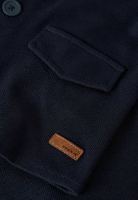 Marine tekstureret stof med en knap og en klaplomme. Har en brun læderlabel med præget tekst "navngiv det."