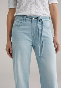 Pantaloni in denim azzurro chiaro a vita alta con cintura annodata, design a gamba larga e tasche frontali. Il tessuto presenta una texture sottile con dettagli di cucitura.