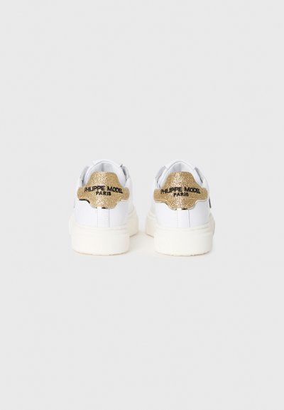 White leather sneakers with textured gold accents on the heel. Features a logo, padded collar, and a thick rubber sole.