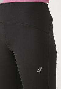 Černé sportovní legíny s hladkou texturou, které mají design švů a malé šedé logo na dolní části nohy.