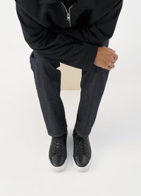 Μαύρα δερμάτινα αθλητικά παπούτσια με λευκή ελαστική σόλα, με στρογγυλές μύτες κα�ι σχέδιο με κορδόνια, συνδυασμένα με σκούρα τζην παντελόνια και μαύρο μπουφάν.