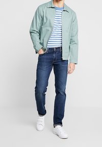 Ljusgrön jacka över en blå och vit randig T-shirt, tillsammans med mörkblå jeans och vita sneakers.