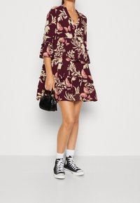 Blommönstrad, vinröd klänning med V-ringning, trekvarts ärmar med vidd, lager av volanger, kombinerad med svarta högstövlade sneakers och en miniväska.