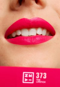 Lys rosa matt leppestift på glatte lepper som avslører hvite tenner. Bakgrunnen viser en lys rosa produktetikett med tekst.