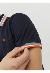 Marineblå polo skjorte med tekstureret stof, orange detaljer på ærmerne og ternet mønster på kraven.