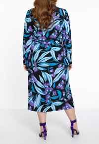 Květinové šaty s wrap stylem, zdobené modrými a fialovými listovými vzory na černém pozadí, dlouhými rukávy a zavazovacím páskem v pase, doplněné o fialové podpatky.