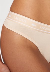 Beige Lacoste stringtruse med en myk, elastisk midje med hvite striper og en liten logo. Glatt tekstur og lavt design.