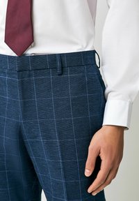 Marineblaue Karohosen mit hellem blauen Rautenmuster, aus strukturiertem Stoff gefertigt. Dazu ein weißes Hemd und eine kastanienbraune Krawatte.