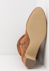 Botte en daim marron avec semelle lisse beige et talon carré bas, positionnée droite contre des murs blancs en angle.