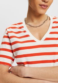 Camiseta de cuello en V en blanco con llamativas rayas horizontales rojas. Tela de mezcla de algodón, mangas cortas y un corte relajado. Collar minimalista de cadena plateada.